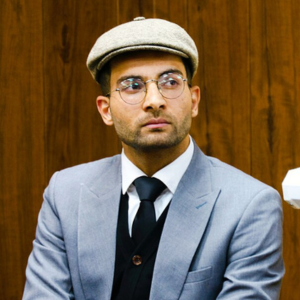 A man wearing a gray suit, black tie, glasses, and a beige flat cap sits in front of a wooden wall, looking slightly to the side with a serious expression.