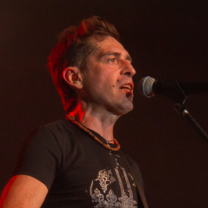A man with short brown hair sings into a microphone on stage. He is wearing a black t-shirt with a white design and a beaded necklace, with dramatic lighting highlighting his face.