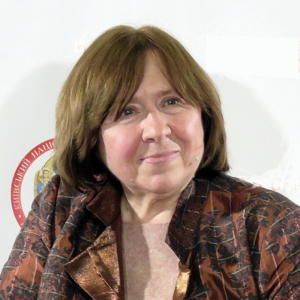A woman with straight, light brown hair and fair skin is wearing a patterned brown jacket and light pink top, posing in front of a white background with red and gold circular text.