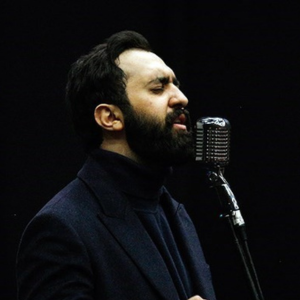 A man with a dark beard and hair, wearing a dark turtleneck and jacket, sings passionately into a vintage-style microphone against a black background.
