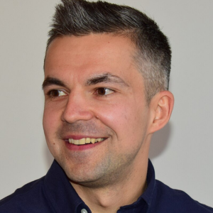 A man with short dark and gray hair, wearing a dark collared shirt, looks to his left and smiles against a plain light-colored background.