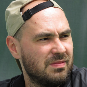 A man with a beard and mustache wears a backward tan cap and a black jacket. He looks slightly to the side with a squint, and the background is blurred and green.