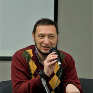 A man with short dark hair and a beard, wearing a red sweater with an argyle pattern, speaks into a microphone while seated indoors. A projector screen is visible in the background.