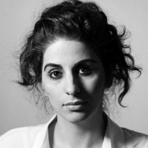 Black and white portrait of a young woman with curly dark hair pulled back, wearing a light-colored top, looking directly at the camera with a neutral expression.