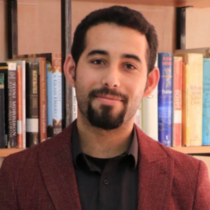 A man with short dark hair and a beard, wearing a maroon blazer and black shirt, stands in front of a bookshelf filled with various books.
