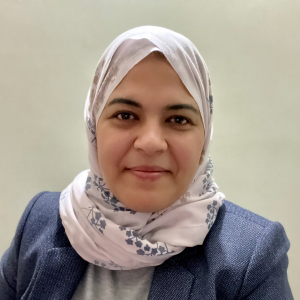 A woman wearing a white floral hijab and a blue blazer smiles at the camera against a plain light background.