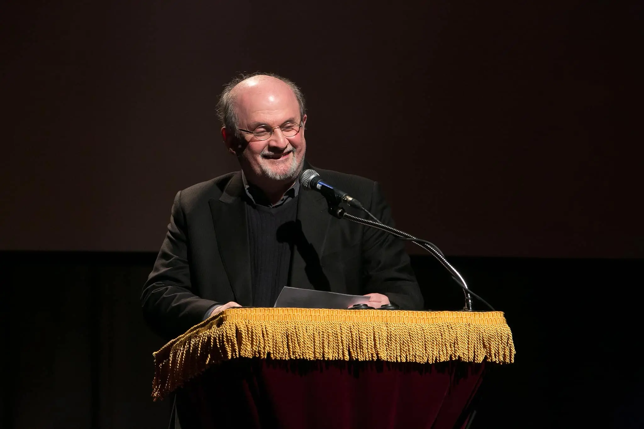 A man with glasses and a beard smiles while speaking at a podium covered with gold fringe, holding a piece of paper and standing in front of a microphone on a dark stage.