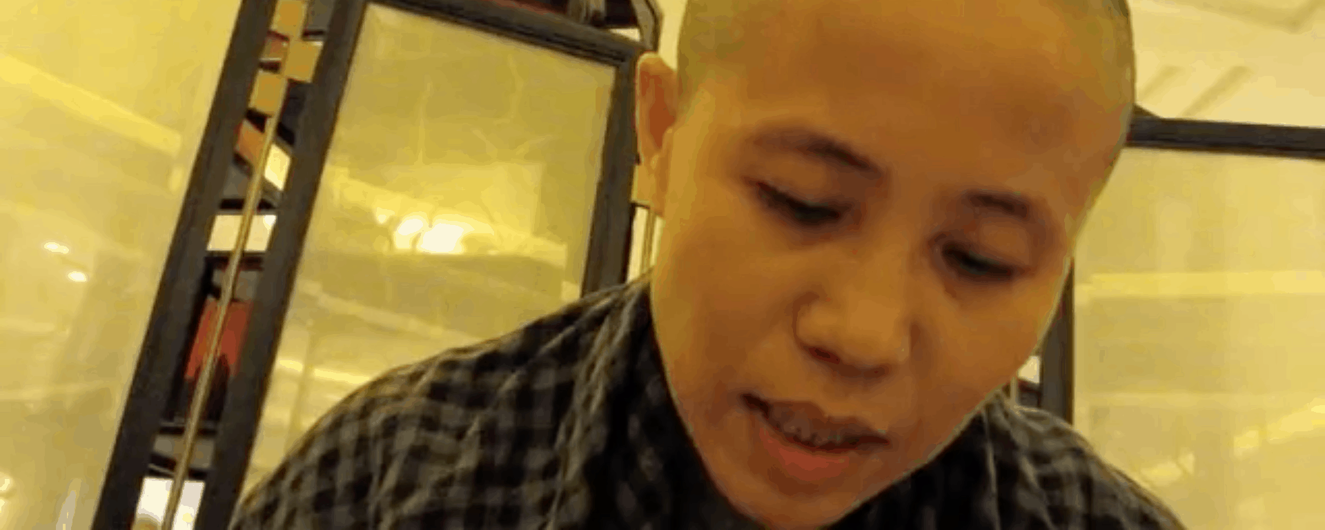 A person with a shaved head and checked shirt is looking down, evoking Liu Xiaobo’s contemplative spirit, with shelves and a partition in the background under warm indoor lighting.