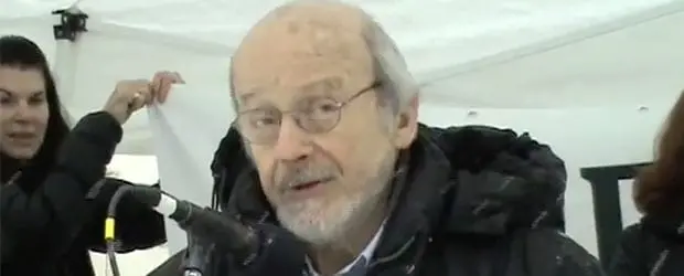 An older man with glasses speaks into a microphone under a white tent, wearing a dark jacket. Two people in the background hold up part of a banner.