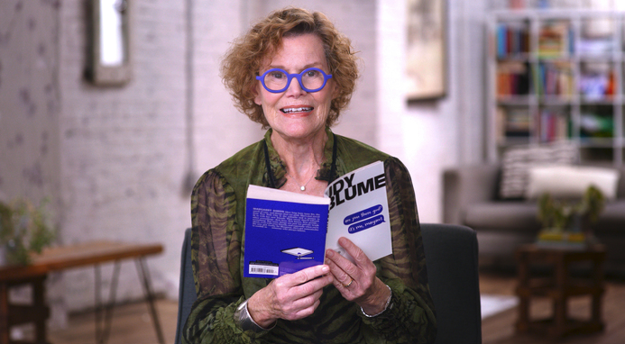 A smiling older woman with curly hair and blue glasses sits indoors, holding and reading a blue and white book. The background shows a cozy, softly lit living room with shelves and a couch.