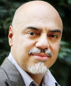 A middle-aged man with a bald head, light skin, a mustache, and a pointed goatee, wearing a striped shirt and a stylish gray blazer, poses confidently outdoors with greenery in the blurred background—your options for refined sophistication.