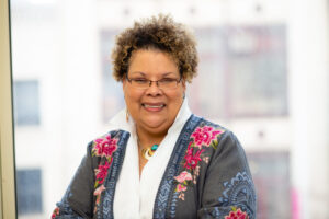A woman with curly hair, glasses, and a warm smile poses indoors. She is wearing a gray jacket with floral embroidery over a white shirt and a statement necklace. The background is softly blurred.