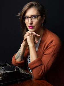 A person with long brown hair, wearing glasses, a rust-colored sweater, and hoop earrings sits at a table with a vintage typewriter, hands clasped, looking thoughtfully toward the camera against a dark background.