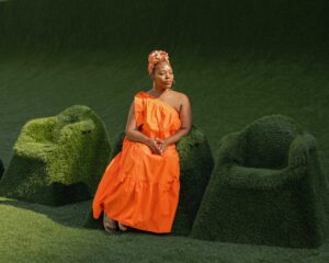A woman in a bright orange, one-shoulder dress and headwrap sits on a green grass-covered chair, with two empty grass-covered chairs beside her, against a matching grassy background.