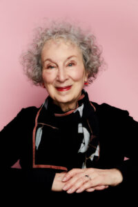 An older woman with curly gray hair smiles warmly, wearing a black top and patterned scarf, posed with her hands folded against a pink background at the Pen America 100 Symposium event.