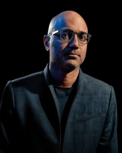 A man with a shaved head, glasses, and a short beard wears a dark jacket over a black shirt. He looks at the camera with a neutral expression against a black background, as if ready to speak at the PEN America 100 Symposium event.