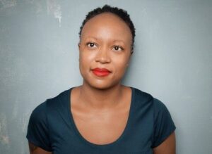 A woman with curly hair wearing large hoop earrings and a floral patterned top stands in front of a dark, plain background, looking confidently at the camera—ready for new year new books in vibrant Los Angeles 2023.