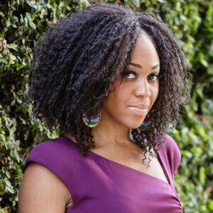 A woman with curly dark hair, wearing a purple dress and colorful beaded earrings, stands in front of a leafy green background in Los Angeles 2023, looking at the camera with a slight smile—ready for new year new books adventures.