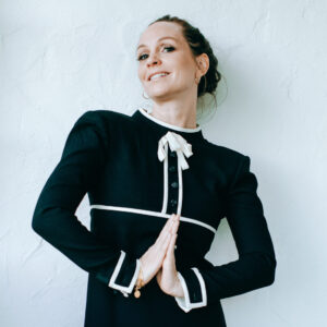 Woman in a black dress with white trim poses against a white wall in Los Angeles, smiling and pressing her palms together. Her hair is tied back and she wears hoop earrings and a bracelet—radiating new year positivity in 2023.