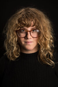 A person with curly, shoulder-length blonde hair wears large round glasses and a black sweater, posing against a plain dark background—capturing the creative spirit of Los Angeles in 2023, ready for new year new books inspiration.