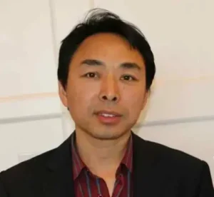 A man with short black hair, wearing a dark blazer over a red striped shirt, stands in front of a light-colored wall at the Liu Xiaobo Vigil.