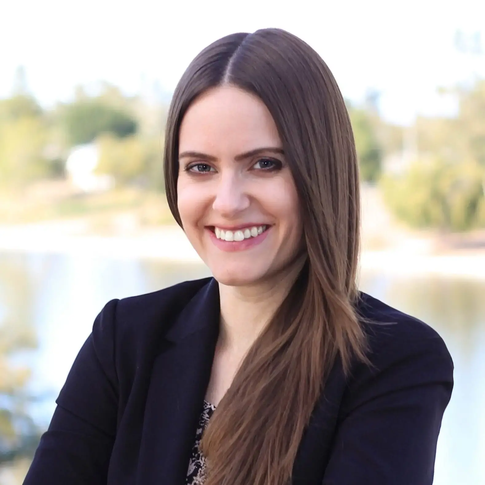 A woman with long brown hair, wearing a black blazer, smiles at the camera outdoors with blurred trees and water in the background—a calm moment reflecting on Free Expression and Schools.