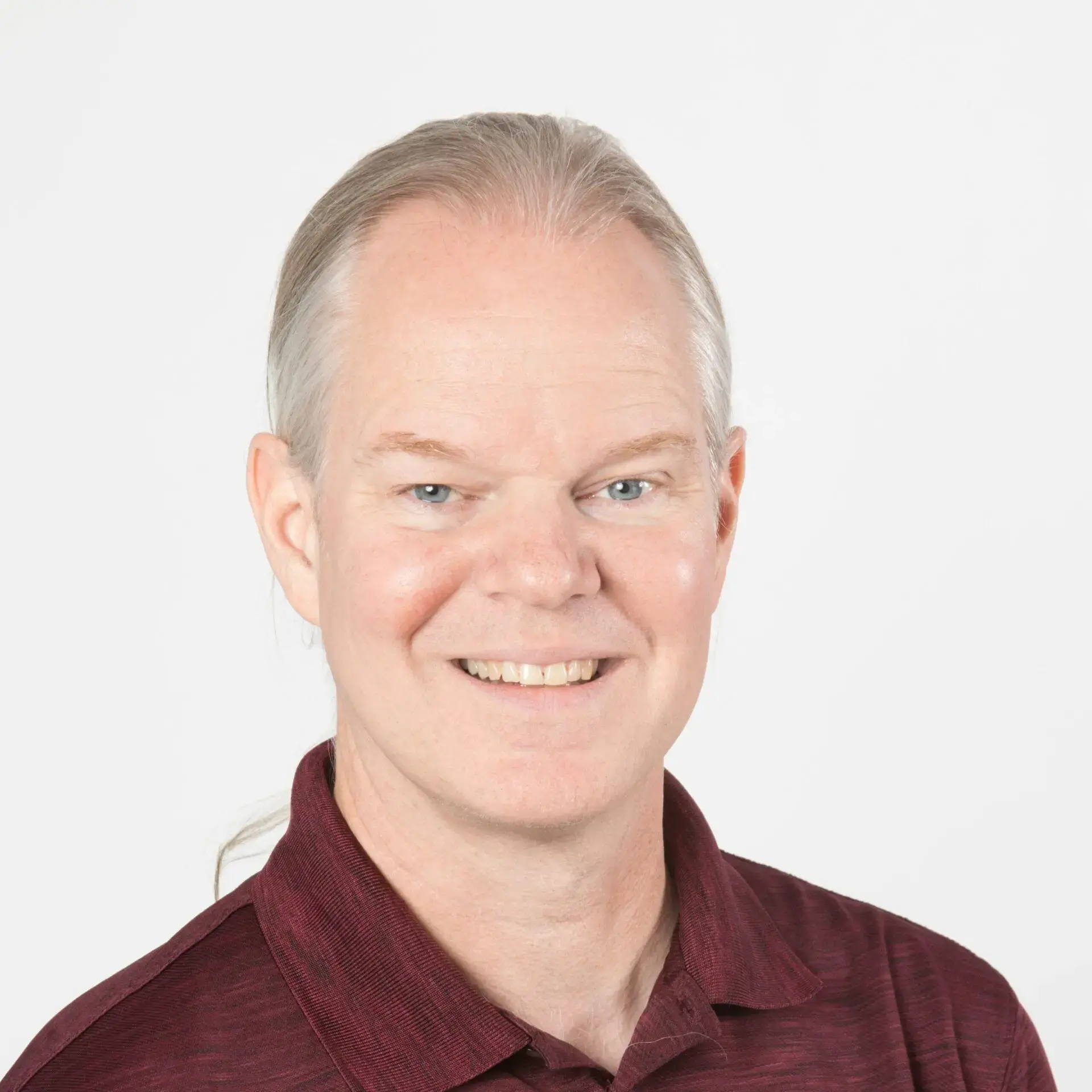 A middle-aged man with light skin, straight gray-blond hair tied back, and blue eyes is smiling at the camera. He wears a maroon collared shirt against a plain white background, embodying themes of free expression in schools.