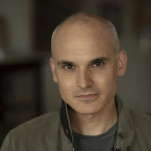 A middle-aged man with a shaved head and brown eyes gazes calmly at the camera, wearing a grey jacket over a dark shirt—his poised demeanor echoing that of a Pulitzer-winning journalist—set against a softly blurred background.