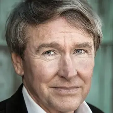 A middle-aged man with light skin, grayish hair, and blue eyes is wearing a white shirt and dark jacket, looking directly at the camera with a slight smile. The softly blurred background suggests an upscale Miami setting.