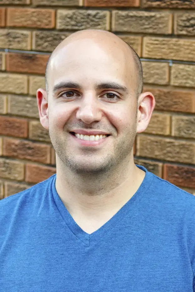 A man with a shaved head and light skin smiles at the camera. He is wearing a blue V-neck shirt and standing in front of a brick wall.
