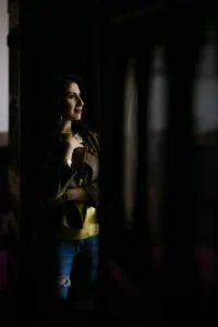 A woman in a brown jacket and ripped jeans stands in a dimly lit room, looking thoughtfully out of a window with her arms crossed and a gentle smile on her face.