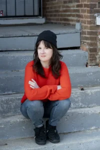 Tracie McMillan, with long brown hair, wearing a black beanie, bright orange sweater, jeans, and black shoes, sits on outdoor concrete steps with arms crossed, looking at the camera.