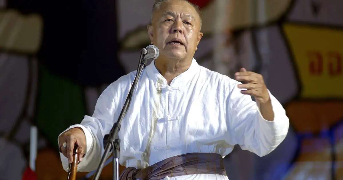 An older man in a white traditional outfit speaks into a microphone on stage, gesturing with one hand and holding a walking stick in the other, with a colorful blurred background behind him.