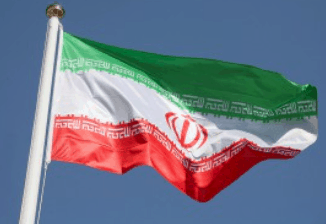 The national flag of Iran waving on a flagpole, featuring horizontal green, white, and red stripes with a red emblem in the center and stylized script along the edges, set against a clear blue sky.
