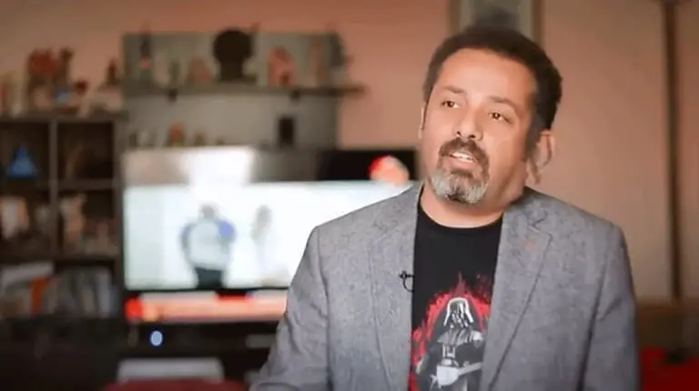 A man with short hair and a beard, wearing a gray blazer and a black Darth Vader t-shirt, sits indoors with shelves and a TV displaying a blurred image in the background.