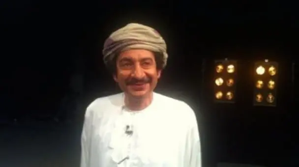 A man in a white traditional robe and a light-colored headscarf smiles at the camera, standing indoors with a dark background and illuminated display behind him.