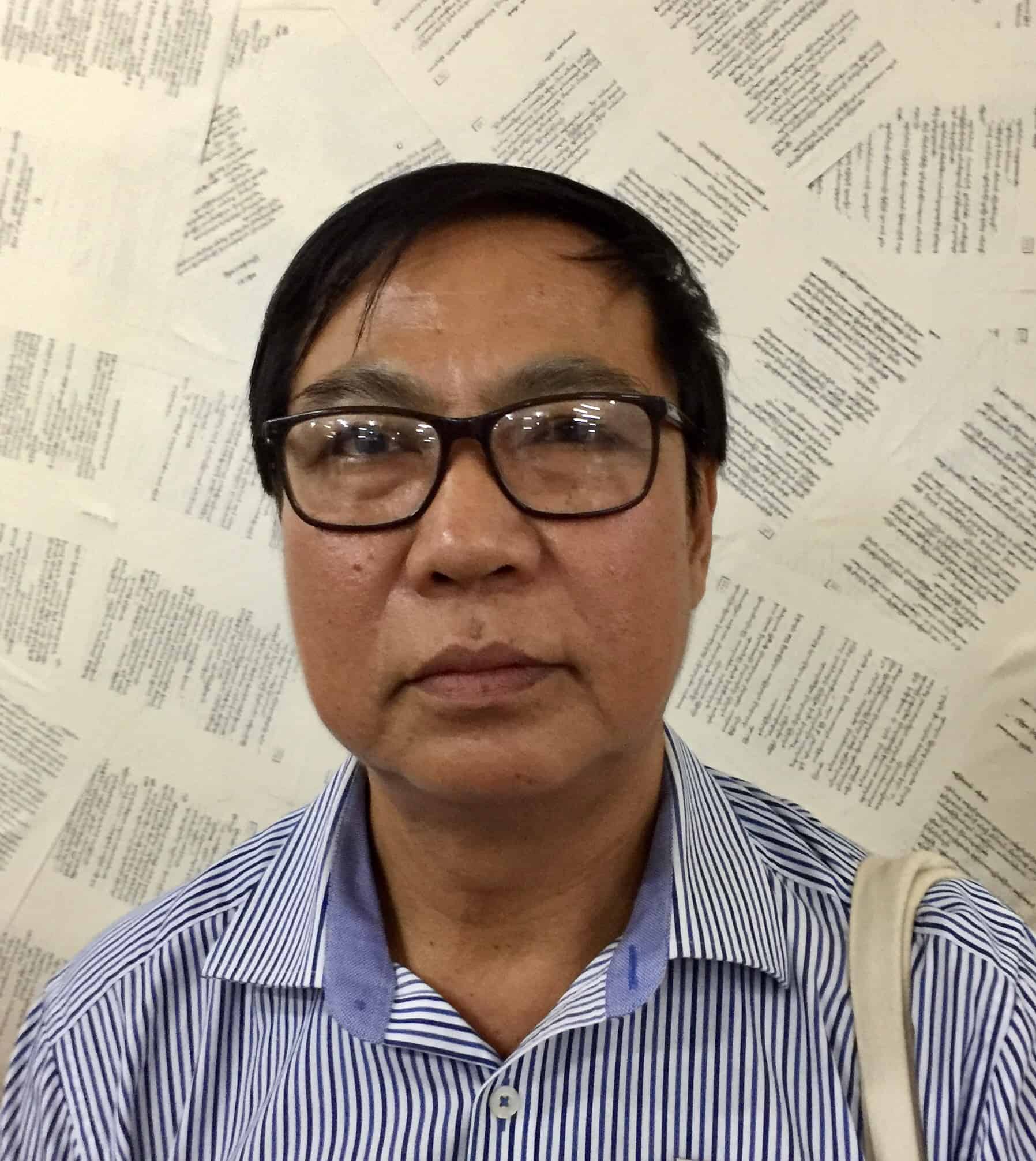 A person with short black hair and glasses wears a blue striped shirt and stands in front of a wall covered with overlapping pages of printed text.