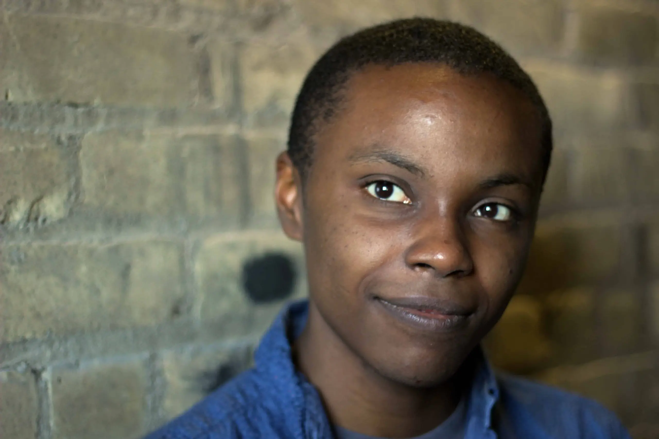 A person with short hair and a blue shirt stands in front of a textured brick wall, looking at the camera with a gentle, slight smile.