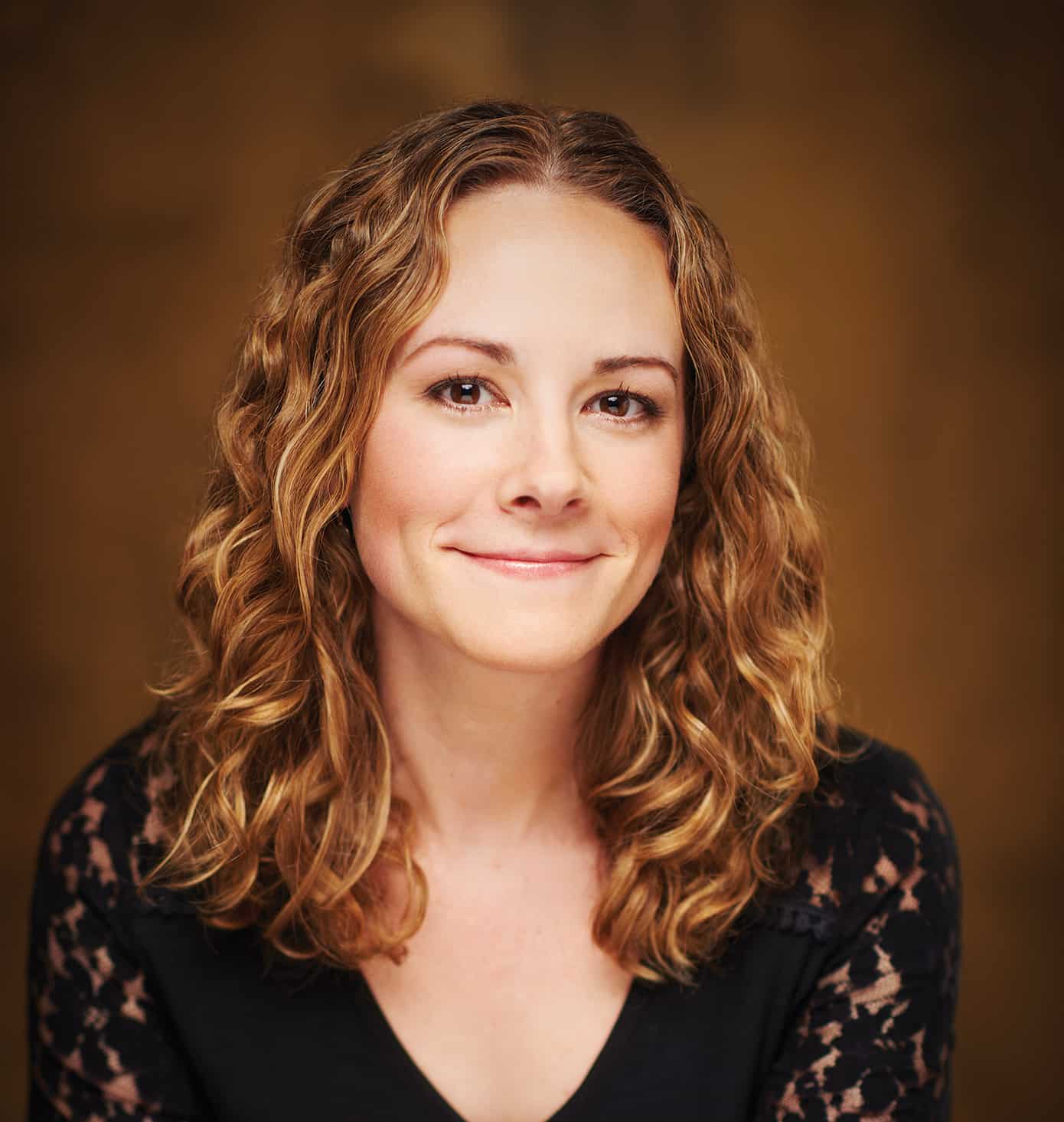 A woman with light brown, curly hair smiles softly at the camera. She is wearing a black top with lace sleeves and is posed against a warm, blurred brown background.