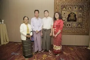 Four people stand indoors in front of a decorative woven tapestry. They are wearing traditional Southeast Asian attire, including long skirts and shirts. Two women and two men face the camera and smile.
