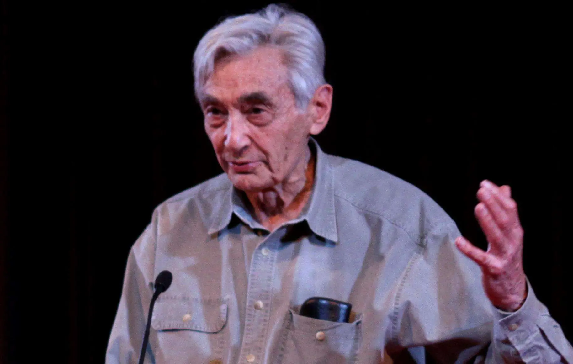 An older man with gray hair speaks at a podium, wearing a light-colored button-up shirt with a pen in the pocket, gesturing with his right hand against a dark background.