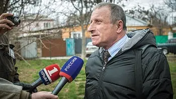 A man in a dark jacket is being interviewed outdoors by reporters holding microphones labeled with Russian news logos. Trees and houses are visible in the background.