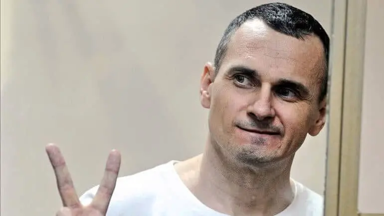 A man with short dark hair wearing a white shirt smiles slightly while making a peace sign with his hand. He is standing indoors near a glass or transparent panel.