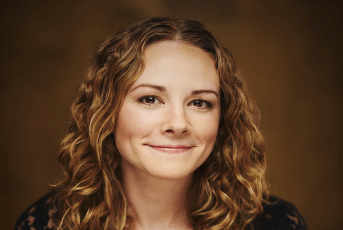 A woman with wavy, light brown hair smiles gently at the camera. She is wearing a dark top and is posed against a soft, brown background.
