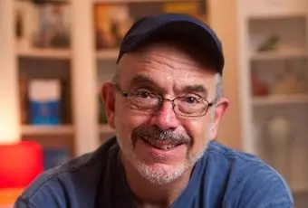 A middle-aged man with glasses, a goatee, and a navy cap smiles warmly at the camera. He is wearing a blue shirt and is indoors, with bookshelves and soft lighting in the background.