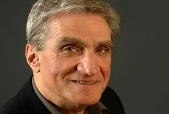 A smiling older man with short gray hair wearing a dark jacket and shirt, pictured against a plain dark background.