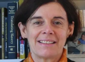 A middle-aged woman with short brown hair smiles in front of a bookshelf containing books, including one titled Translating Slavery. She is wearing an orange scarf and a dark top.