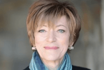 A woman with short, light brown hair and blue eyes smiles softly, wearing a blue scarf and pearl earrings, against a blurred background.