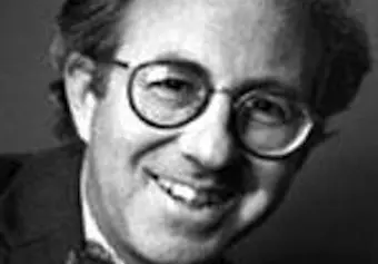 Black and white photo of a smiling person wearing glasses and a suit, with short, wavy hair. The image is slightly blurred.