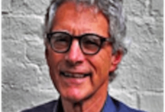 A man with gray curly hair and glasses is smiling in front of a textured light-colored brick wall. He is wearing a dark jacket and a collared shirt.